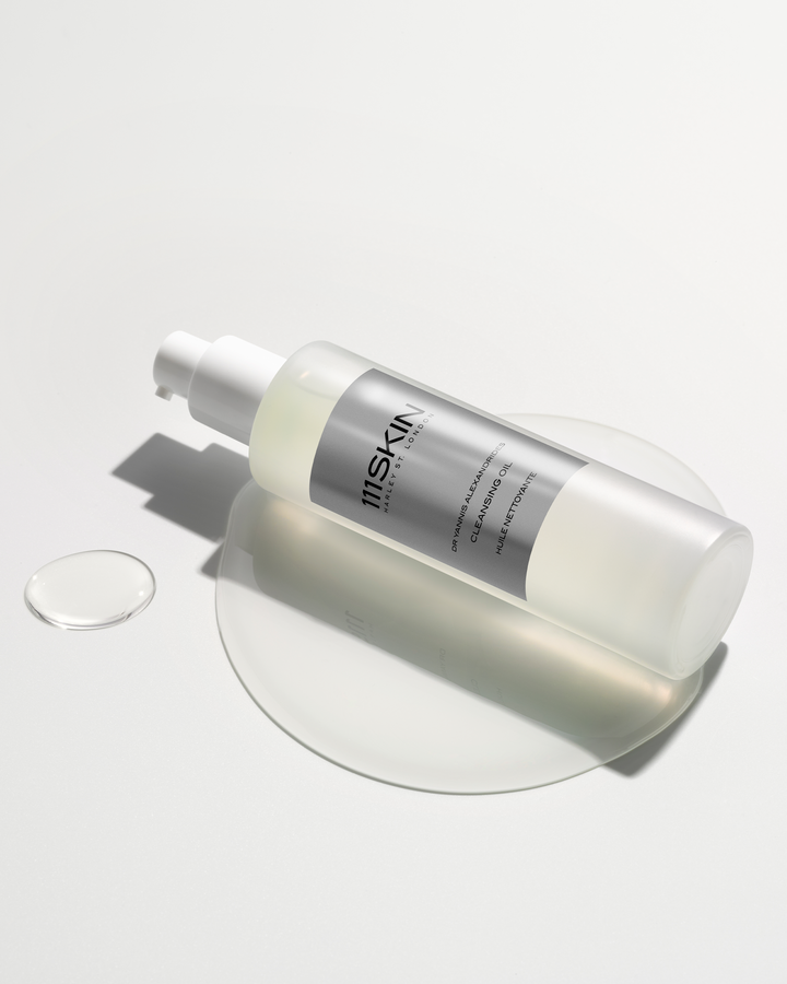 A 111SKIN cleansing oil bottle resting on a white surface amidst a puddle and a droplet of clear oil.