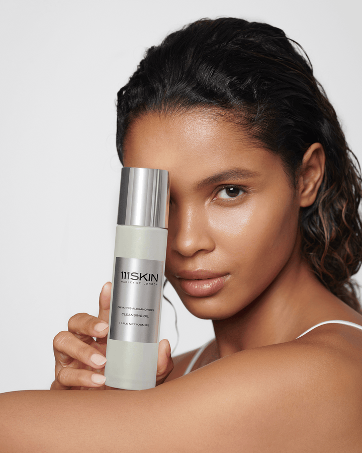 A woman holding a bottle of 111SKIN Cleansing Oil vertically over one eye.