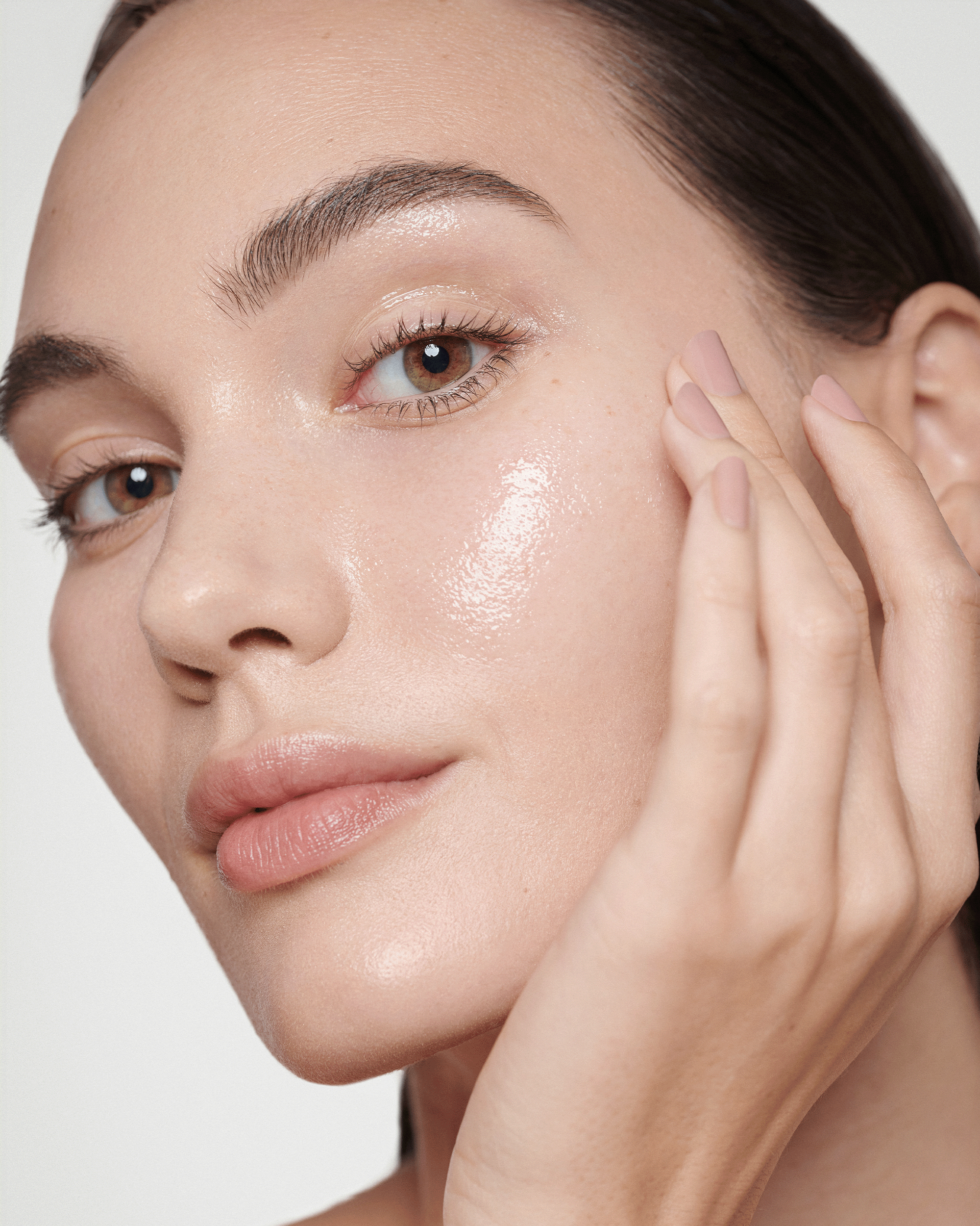 A close-up profile view of a woman with dewy, radiant skin touching her cheek with her hand.