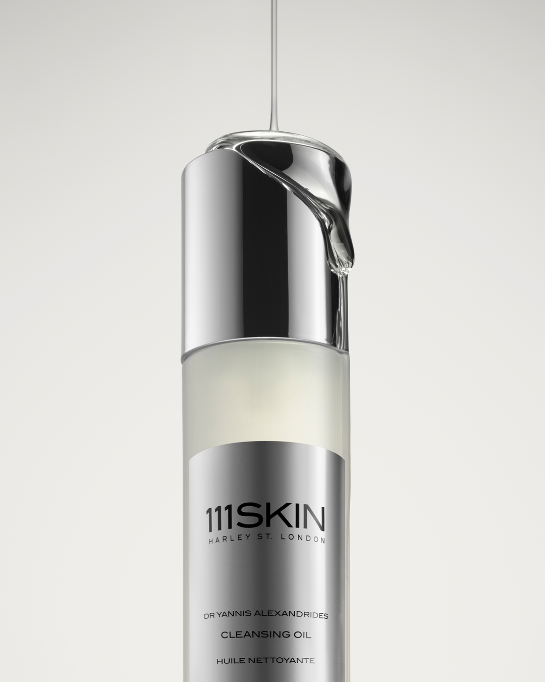 A stream of clear, viscous oil pouring directly onto the reflective silver cap of a 111SKIN Cleansing Oil bottle.
