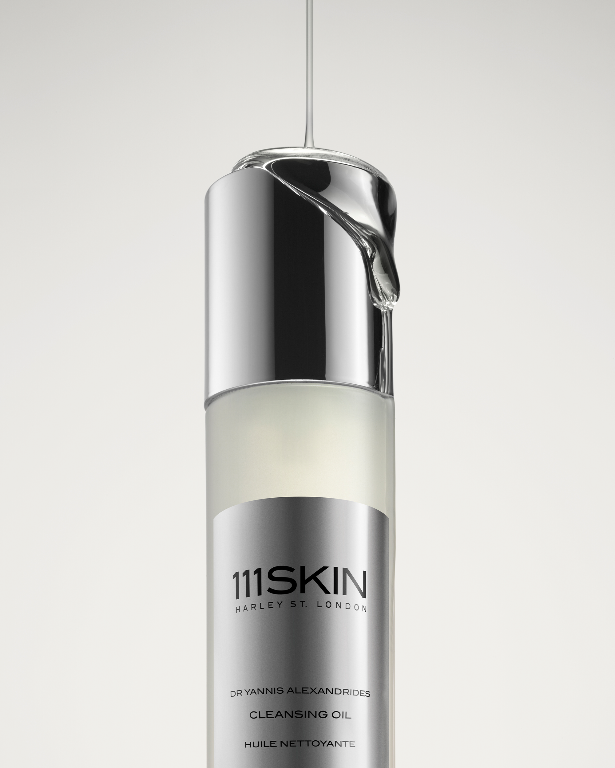 A stream of clear, viscous oil pouring directly onto the reflective silver cap of a 111SKIN Cleansing Oil bottle.