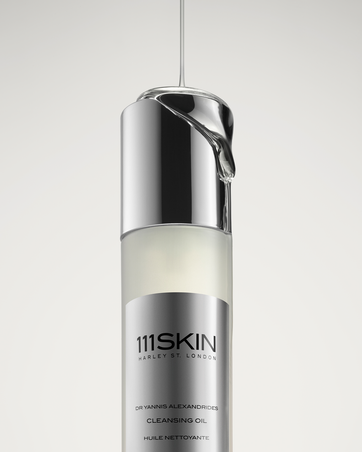 A stream of clear, viscous oil pouring directly onto the reflective silver cap of a 111SKIN Cleansing Oil bottle.