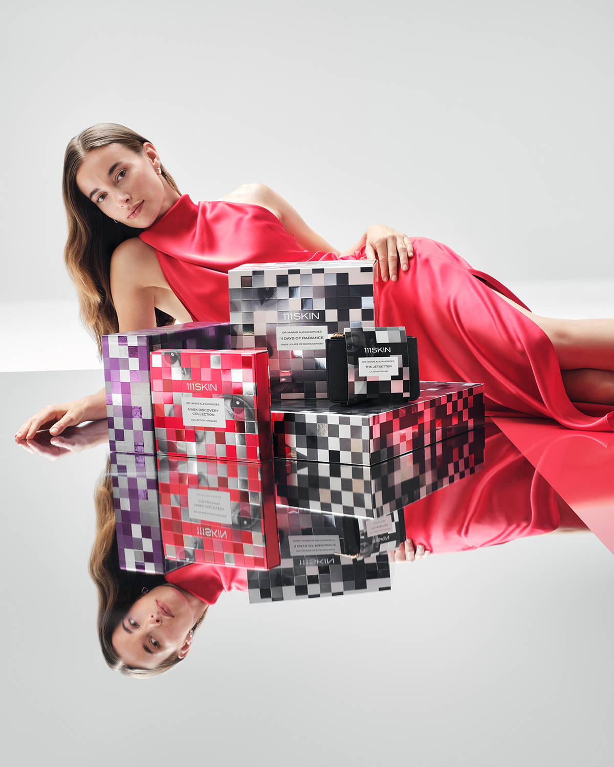 Model in a red satin dress posed beside 111SKIN holiday gift sets, including the Mask Discovery Collection, 11 Days of Radiance, and The Jetsetter.