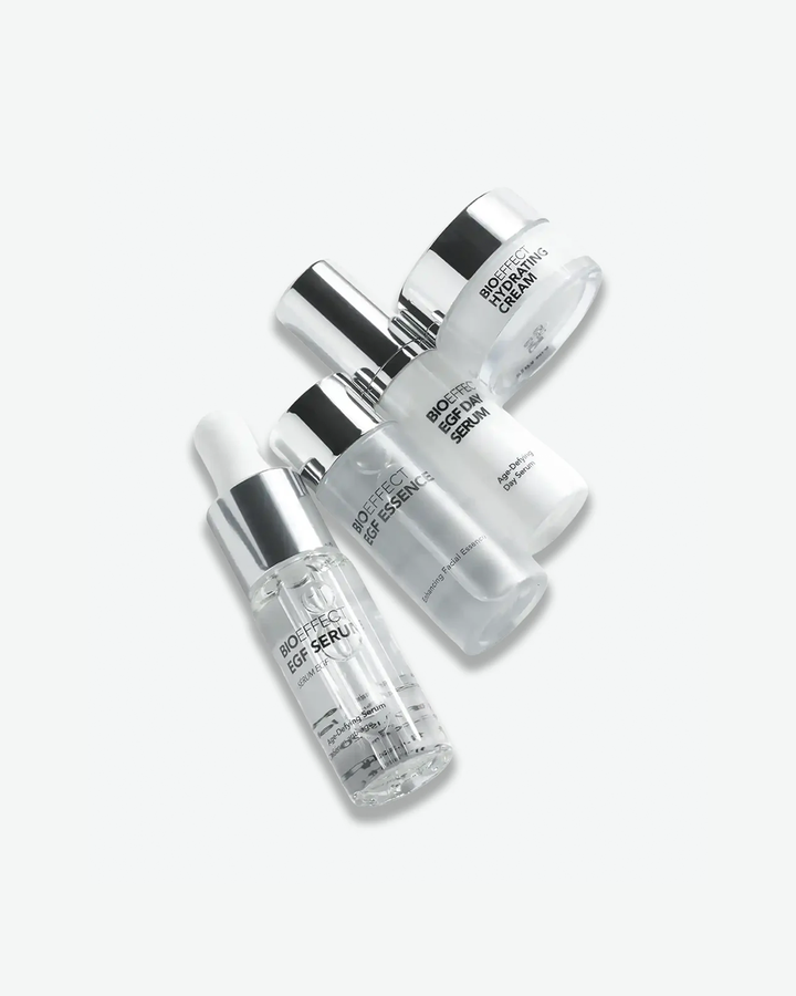 A close-up, top-down shot of four Bioeffect skincare products arranged diagonally. The products include a clear glass dropper bottle of Bioeffect EGF Serum, a frosted glass bottle of Bioeffect EGF Essence, a white bottle of Bioeffect EGF Day Serum, and a small glass jar of Bioeffect Hydrating Cream, all featuring metallic silver caps.