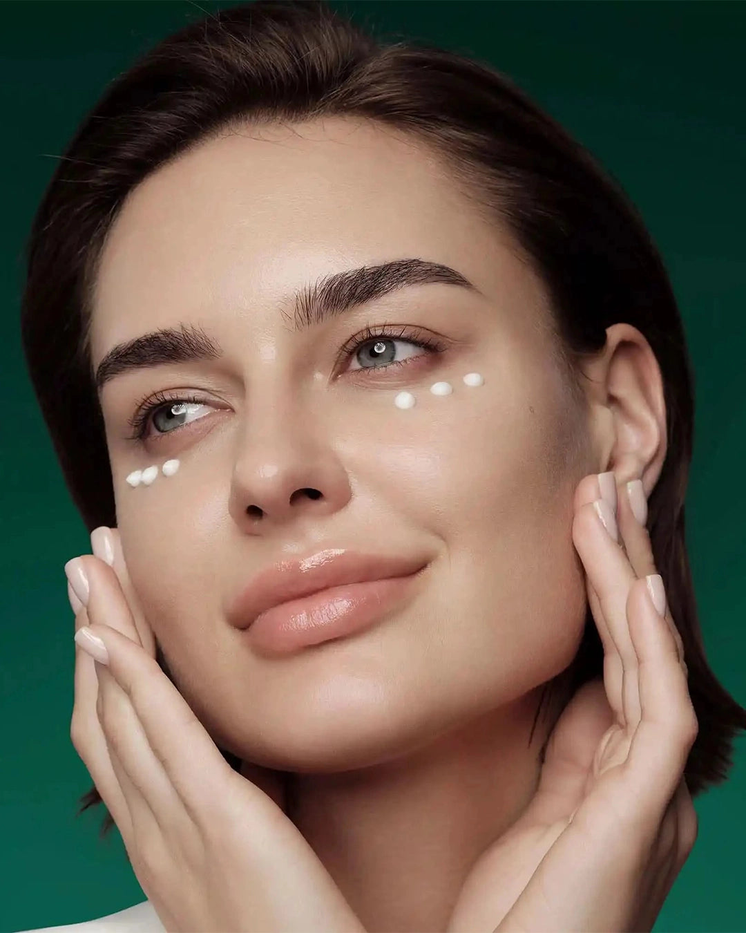 A close-up shot of a model with short brown hair against a dark green background. She is smiling gently with her hands cupping her face, showing three small dots of white eye cream applied neatly under each eye.