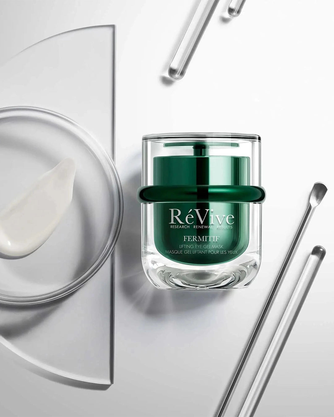 An artistic, high-angle flat lay featuring the RéVive eye gel mask jar resting on a clean white surface alongside glass rods, a silver cosmetic spatula, and a clear petri dish holding a smooth swatch of the white cream.
