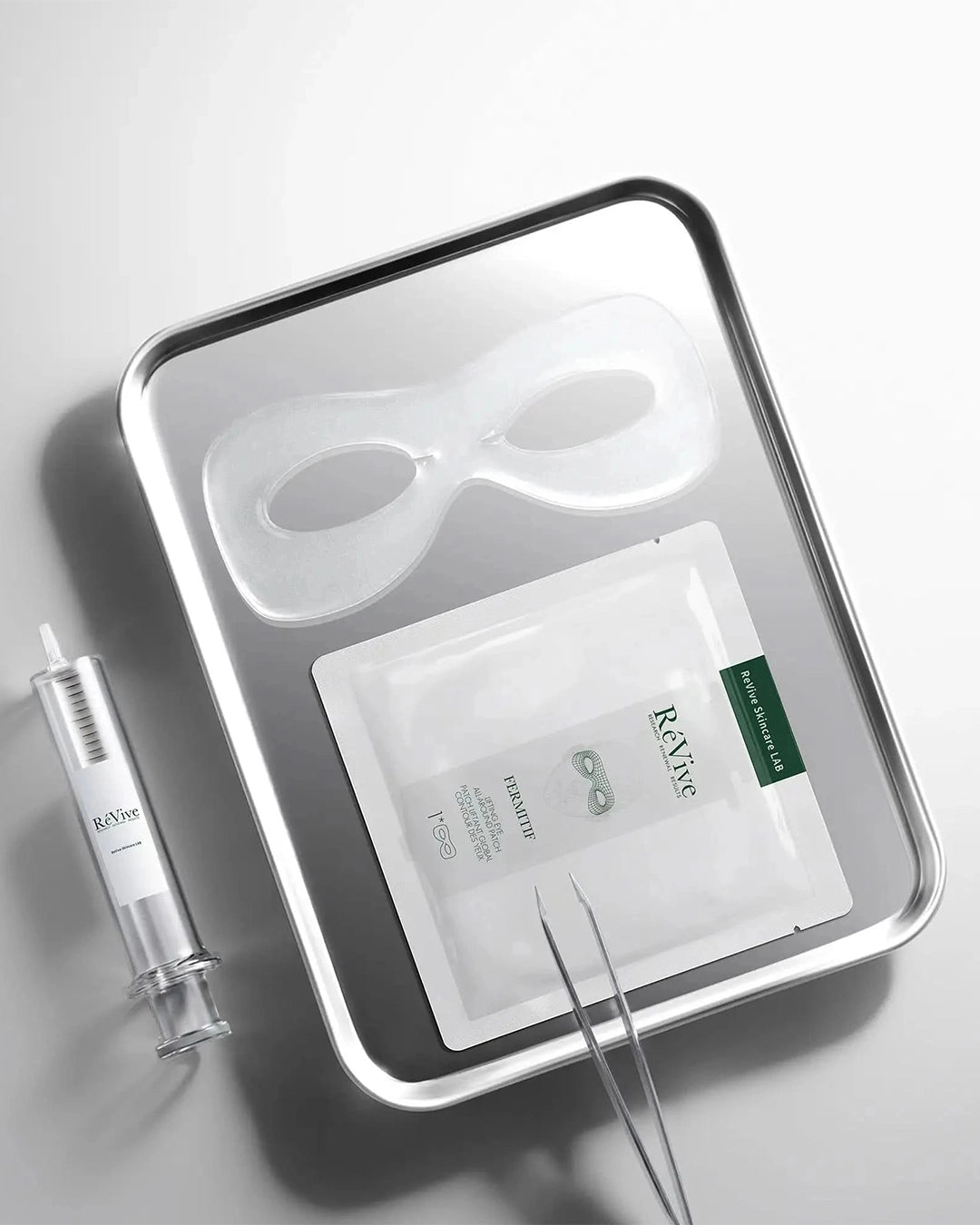 A clinical flat lay on a metallic tray featuring a clear, gel-like eye mask with two eye cutouts, alongside its corresponding RéVive sachet, a clear syringe-like applicator, and metal tweezers.