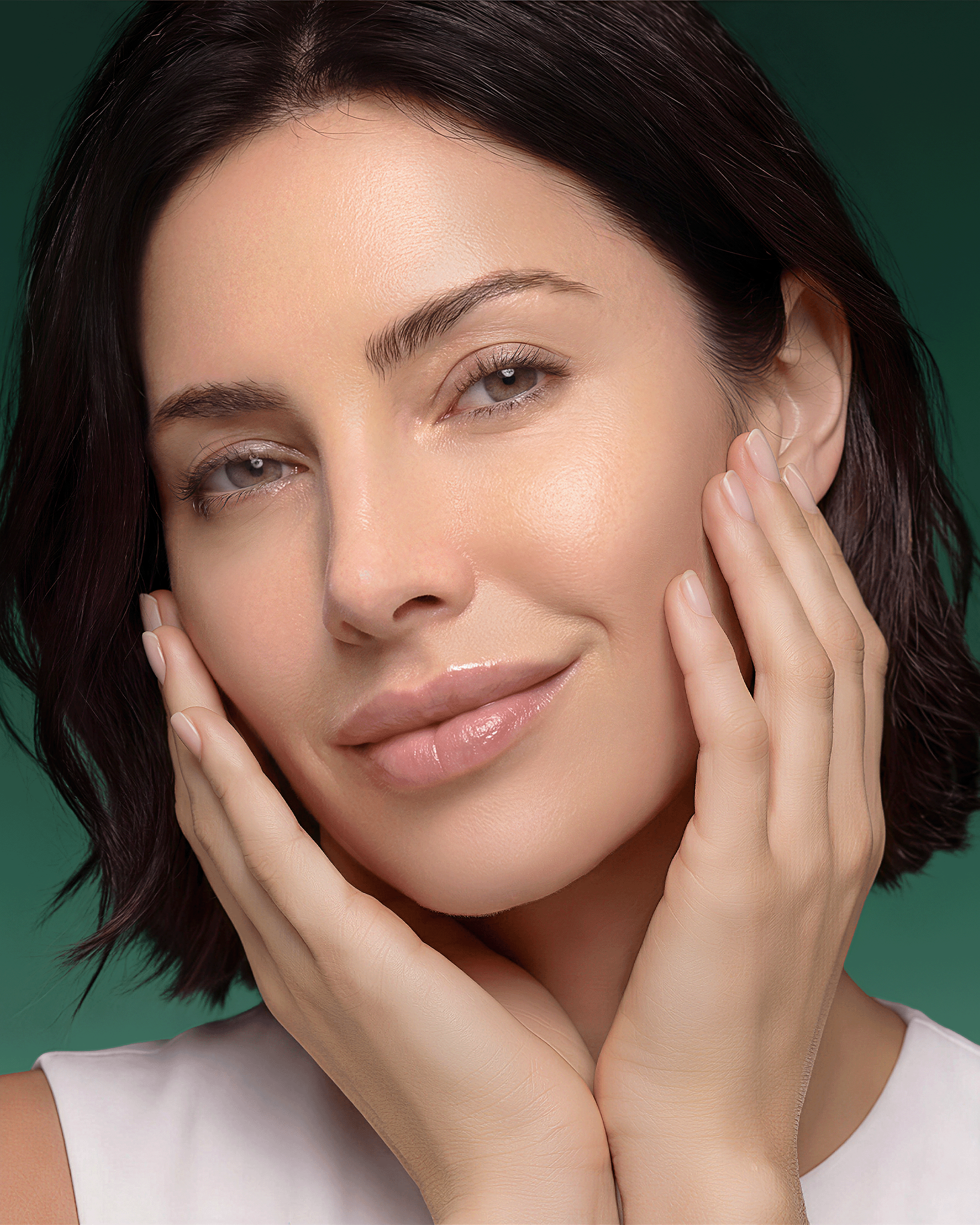 A close-up portrait of a woman with smooth, radiant skin touching her cheeks, set against a solid dark green background.