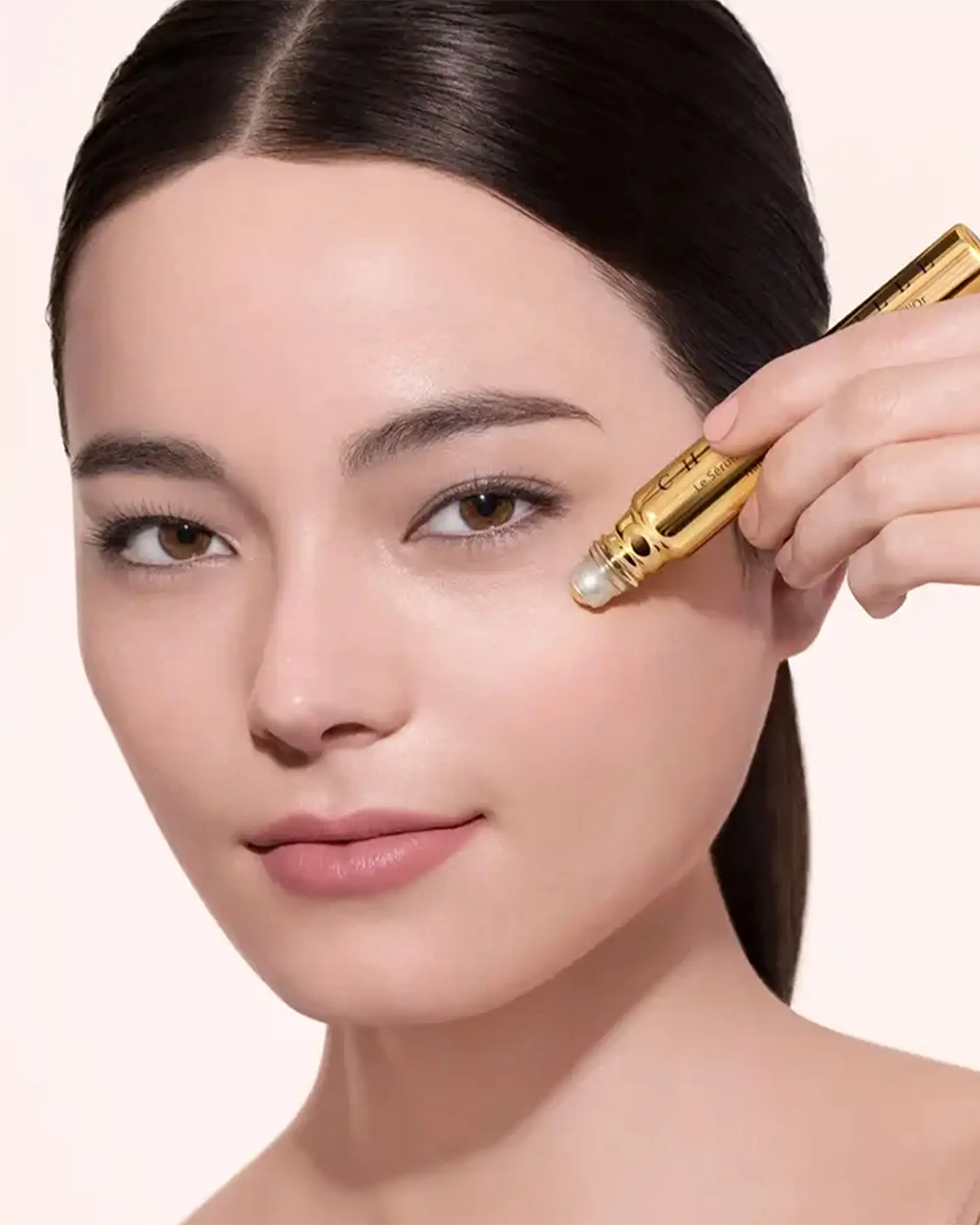 A close-up shot of a woman with dark hair tied back, demonstrating the product by gently gliding the gold Chantecaille rollerball eye serum under her left eye.