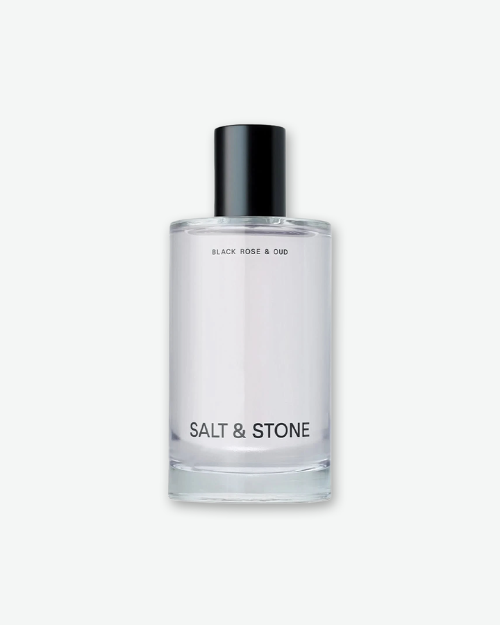 A studio product shot of Salt & Stone Black Rose & Oud Body Mist. The minimalist clear glass cylindrical bottle features a black atomiser cap, with 'BLACK ROSE & OUD' printed at the top and 'SALT & STONE' in bold black text at the bottom.