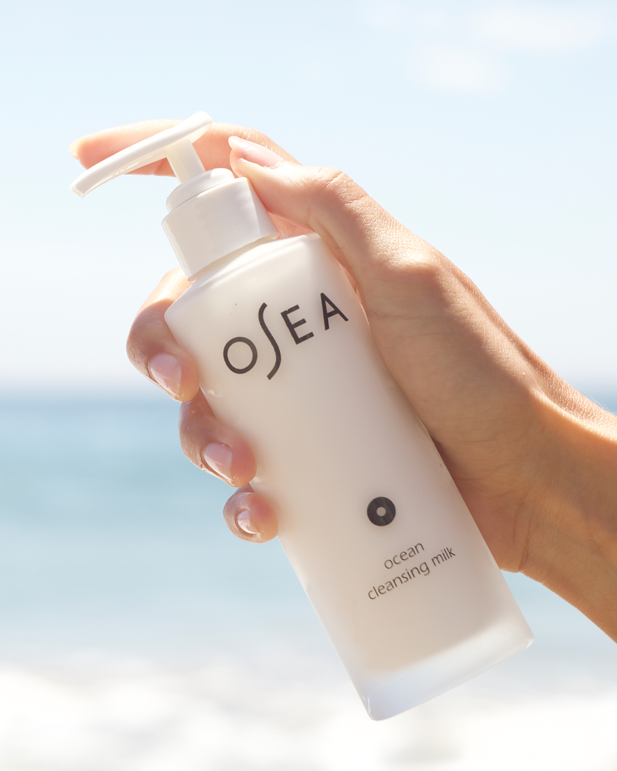 A hand holding a pump bottle of OSEA Ocean Cleansing Milk against a sunny, blurred beach background.