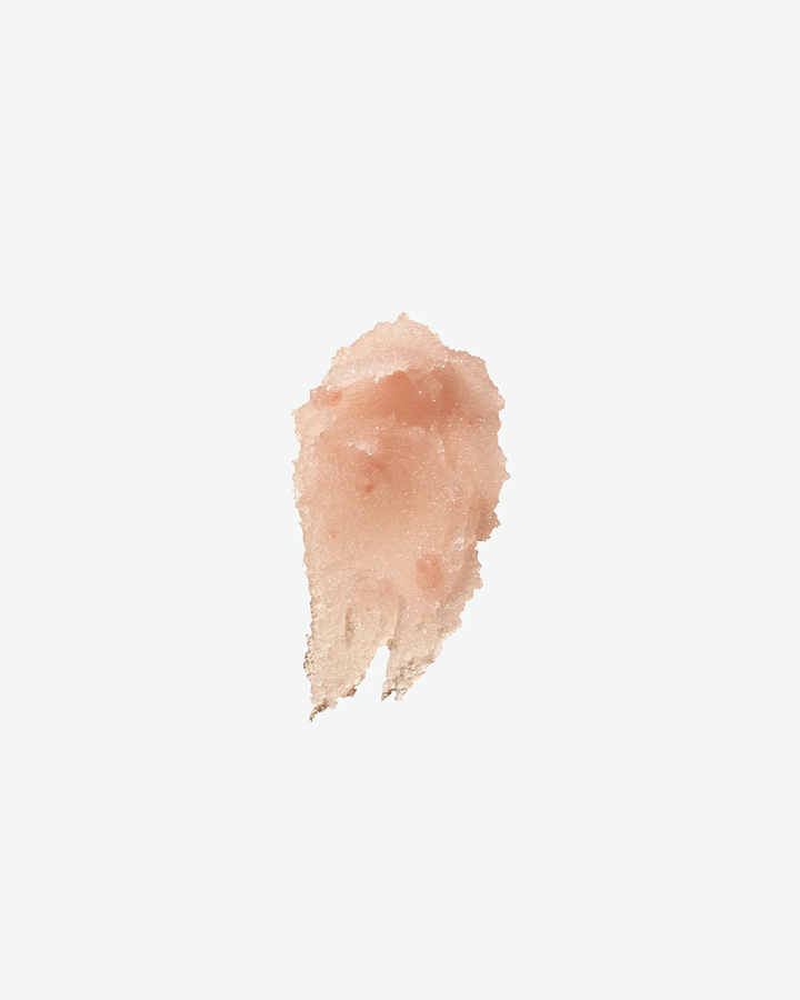 A product texture shot showcasing a textured, grainy smear of pale pink body scrub isolated in the center of a solid white background.