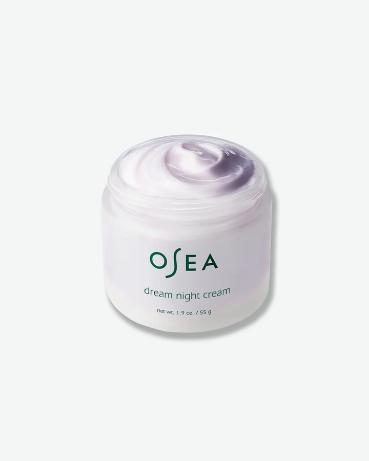 An angled, open-container shot of an OSEA Dream Night Cream jar with the lid removed. The wide, frosted glass jar is filled with a thick, glossy night cream that features a soft white and lavender-purple swirled texture. The dark green OSEA logo and product details are printed on the front of the jar against a plain white background.