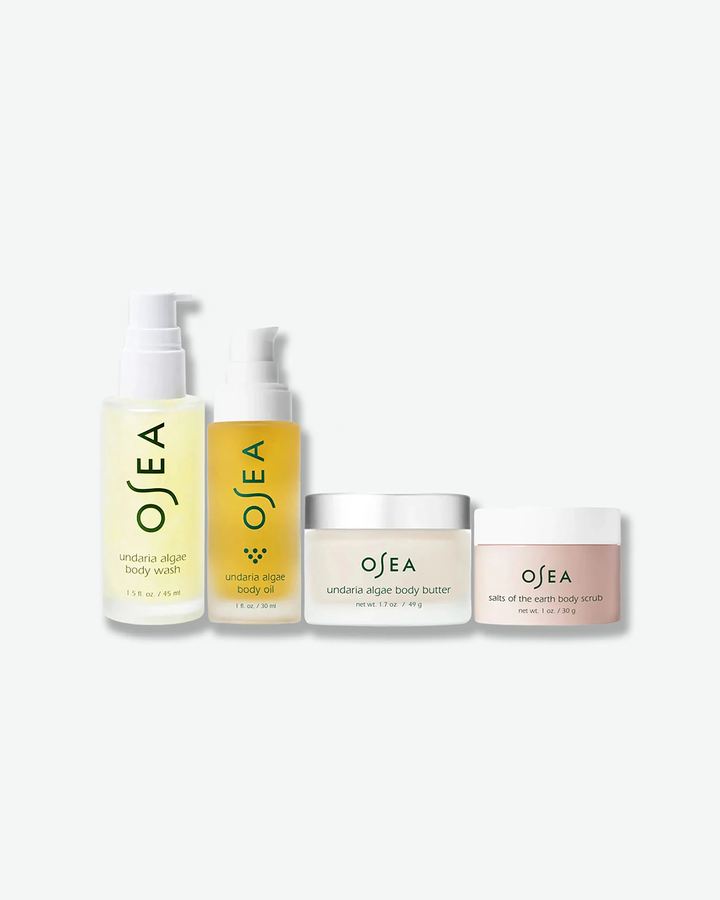 A product group shot showcasing four OSEA travel-sized items arranged by height on a white surface: a translucent yellow body wash, a golden body oil bottle, a clear jar of body butter, and a frosted pink jar of salts of the earth body scrub.