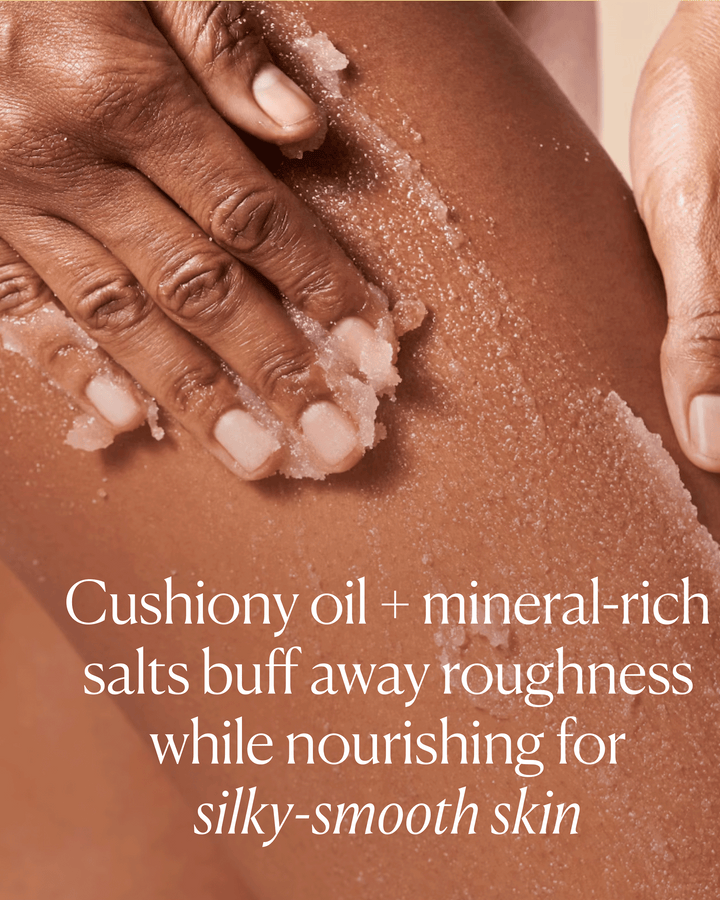 A hand applying a gritty pink body scrub onto skin with text highlighting a "cushiony oil + mineral-rich salts" formula.