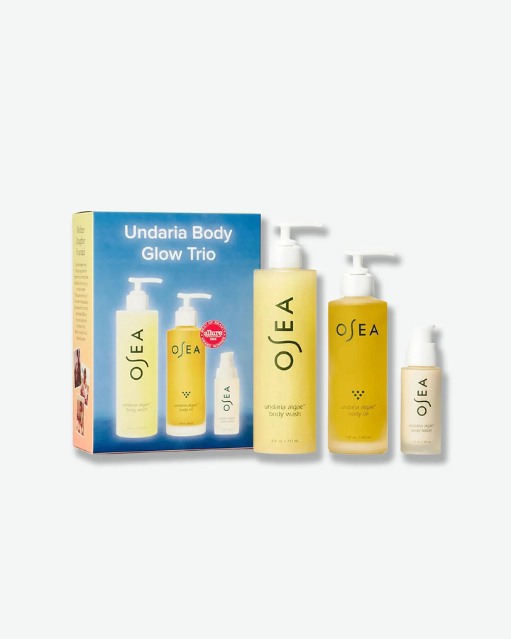 A product kit shot highlighting the OSEA "Undaria Body Glow Trio" box. The gradient blue-to-orange background box stands next to its included three full-size and travel-size golden-hued Undaria algae body products, each fitted with white dispensers.
