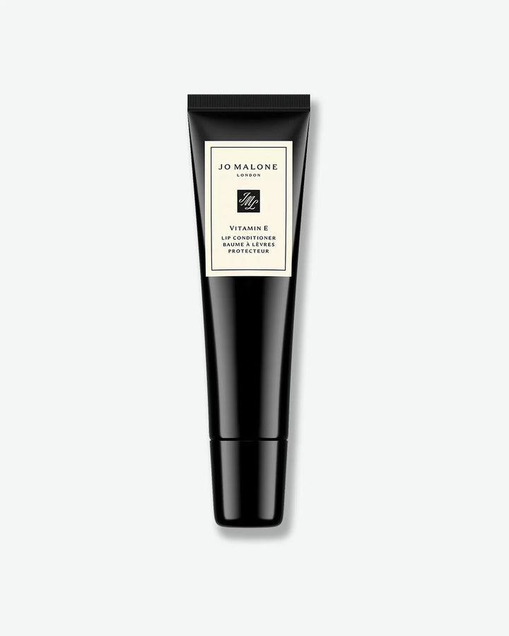 A black squeeze tube labeled “Jo Malone London Vitamin E Lip Conditioner.” The tube features the brand’s signature cream-colored label with black trim and is designed for lip care and protection. Displayed against a light gray background.