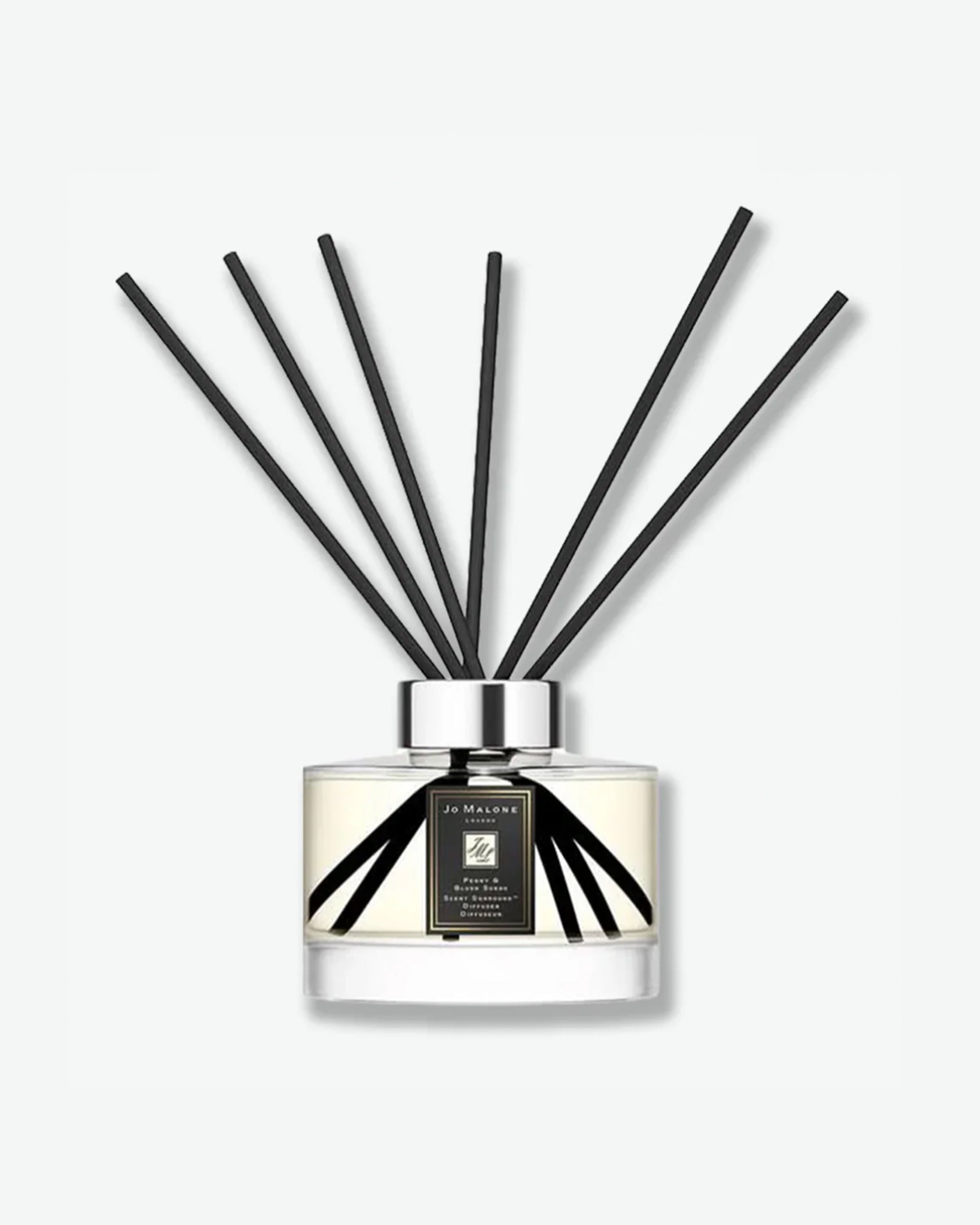 Product shot of a Jo Malone London Peony & Blush Suede reed diffuser. The clear, cylindrical glass bottle is filled with light-colored liquid and features a square black-and-gold label. Six long, black reeds fan out dramatically from the polished silver collar, casting a soft shadow against a clean, minimal white background.