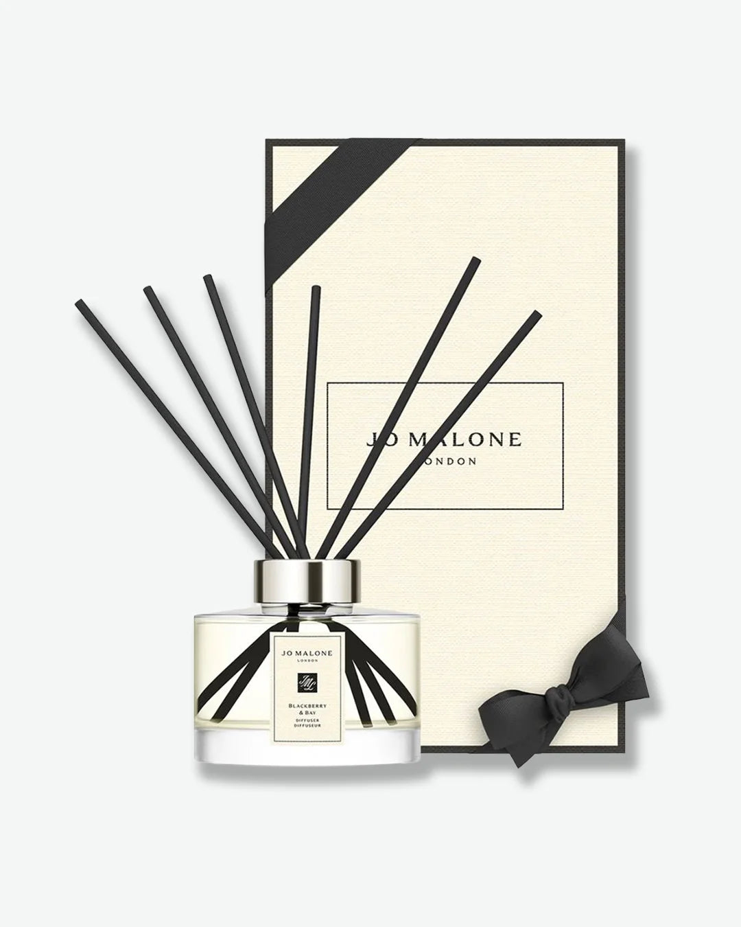 Jo Malone glass jar diffuser with black reeds stood slightly in front of the signature Jo Malone London cream colored packaging box with black ribbon on a white background