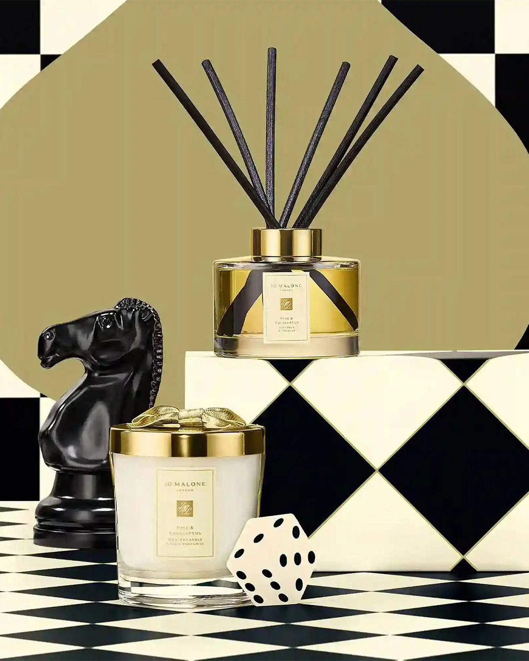 Holiday campaign imagery for Jo Malone London featuring a Pine & Eucalyptus scented candle with a metallic gold lid and a matching reed diffuser with gold trim. The items are arranged on a black-and-white checkered harlequin floor with a black chess knight piece and a white die with black dots. The background displays an olive-green circle on a checkered pattern.
