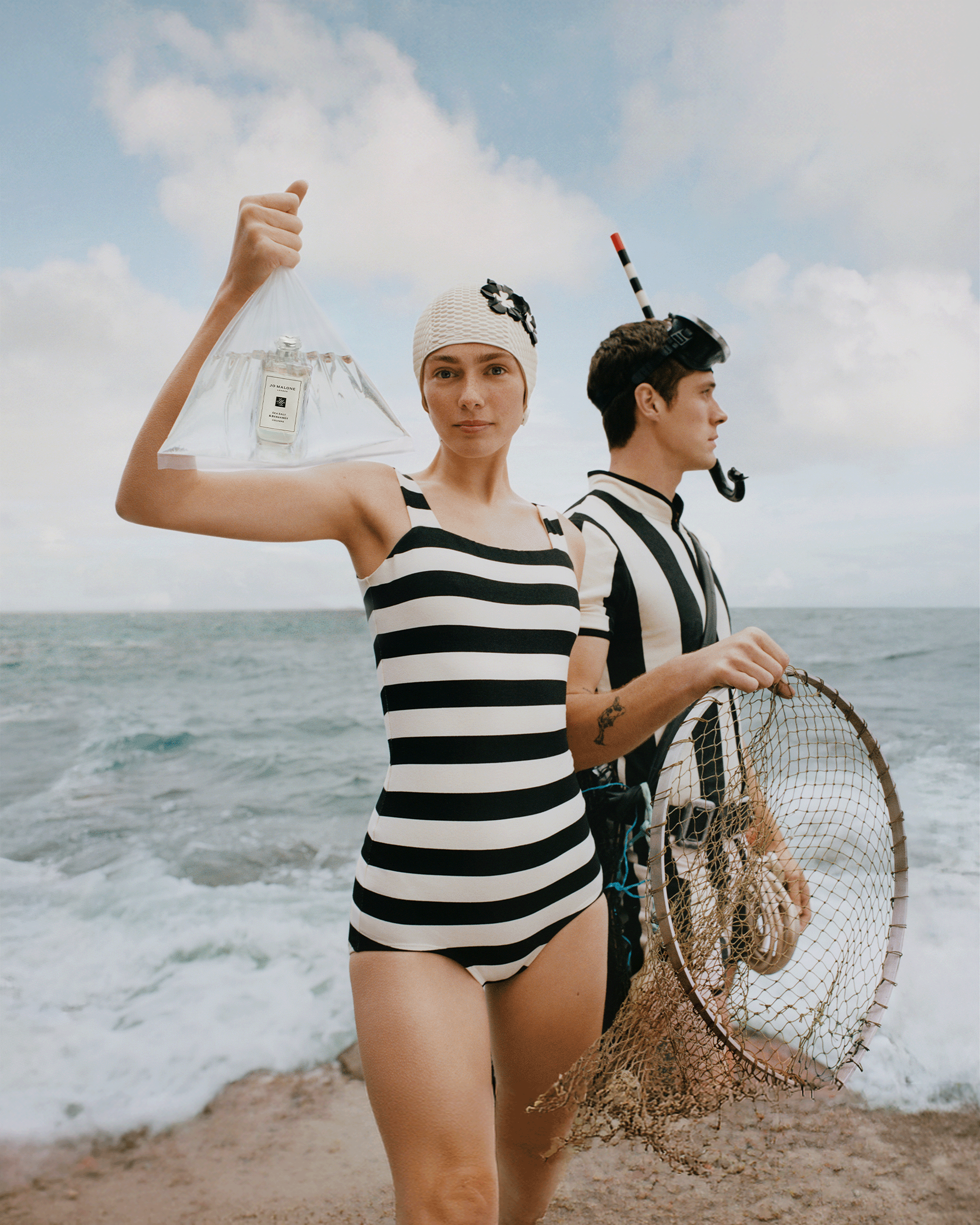 A woman and a man standing on a beach in front of the ocean, both wearing black-and-white striped outfits. The woman holds a clear plastic bag containing a small perfume bottle, and the man holds a fishing net.