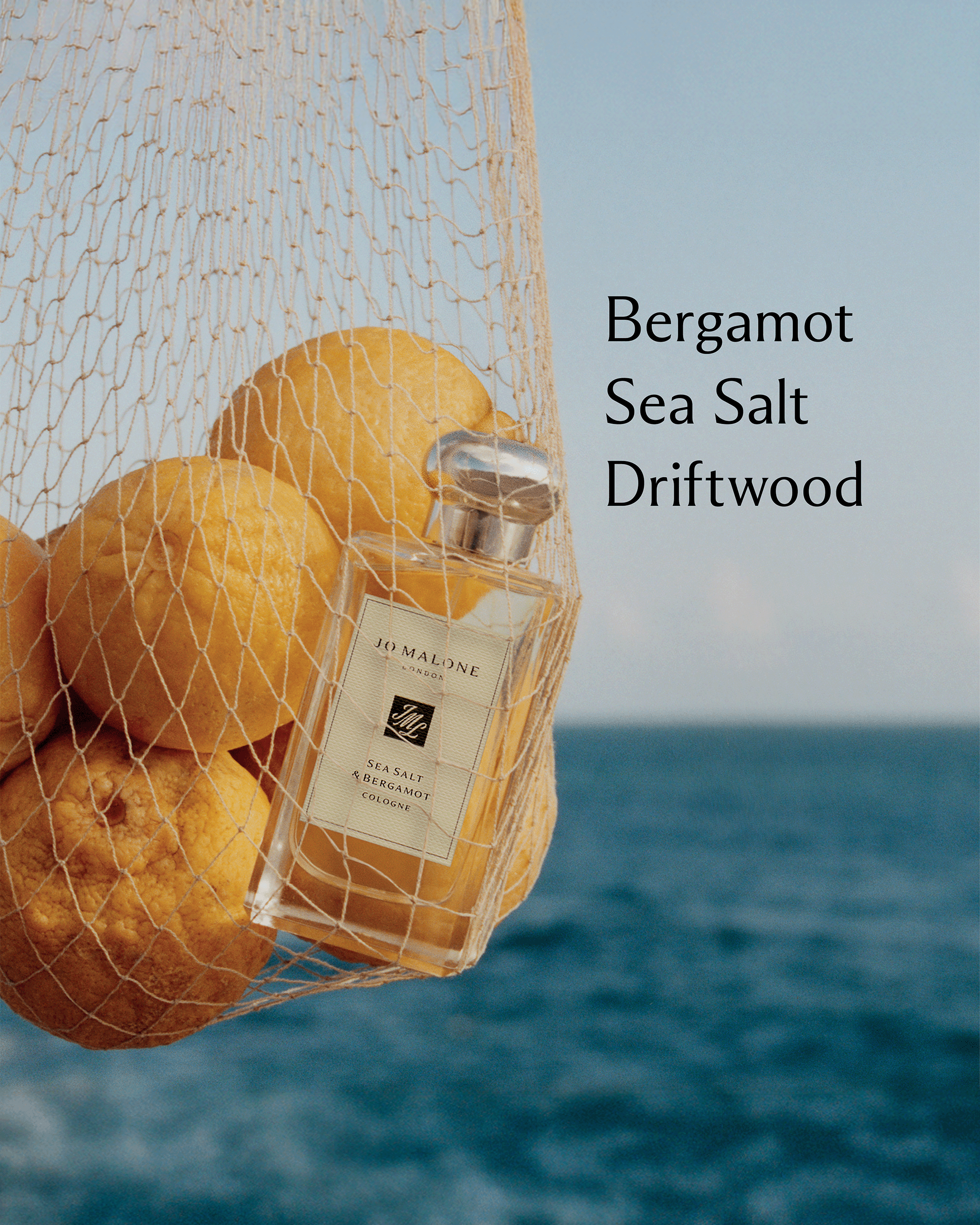 A bottle of Jo Malone London Sea Salt Driftwood perfume hangs in a white mesh bag filled with lemons against a blue ocean and sky background, with the text 'Bergamot Sea Salt Driftwood' on the right.