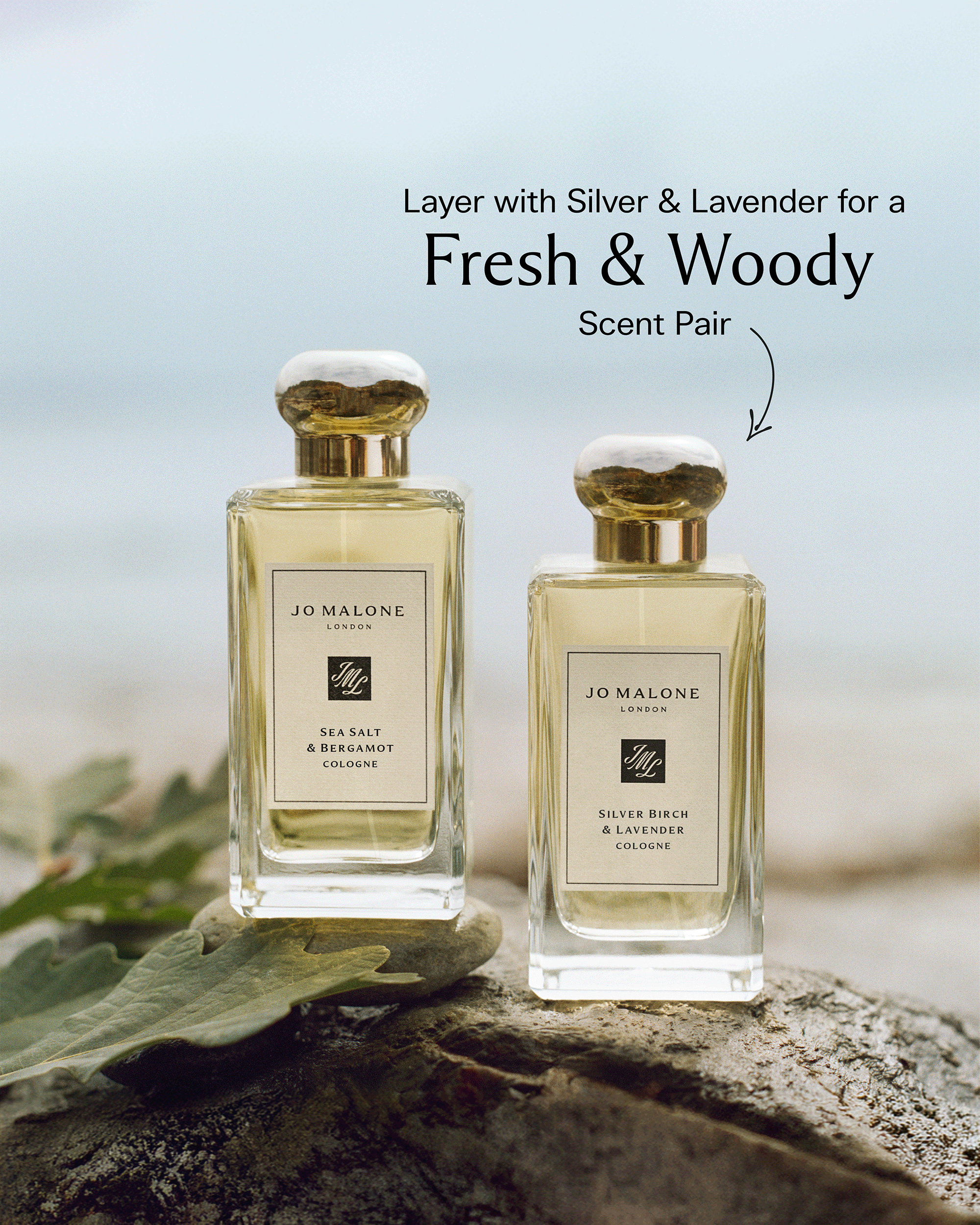 Two JO MALONE London cologne bottles, Sea Salt & Bergamot and Silver Birch & Lavender, displayed on a rock with a blurred beach background. Text overlay reads 'Layer with Silver & Lavender for a Fresh & Woody Scent Pair'.