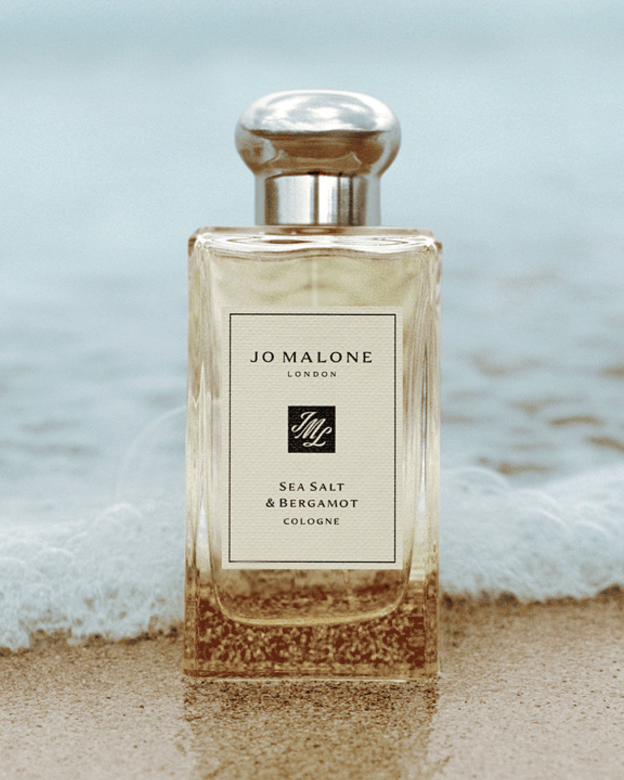 A bottle of Jo Malone London Sea Salt & Bergamot Cologne stands on wet sand with ocean waves blurred in the background. The clear glass bottle shows a warm beige liquid and a white label with black text and logo.