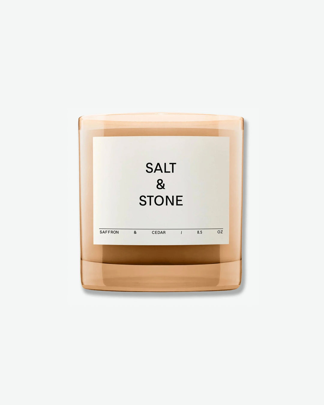 Salt & Stone Saffron & Cedar 8.5 oz scented candle in a minimalist translucent amber glass jar with a square off-white label, isolated against a light grey background.