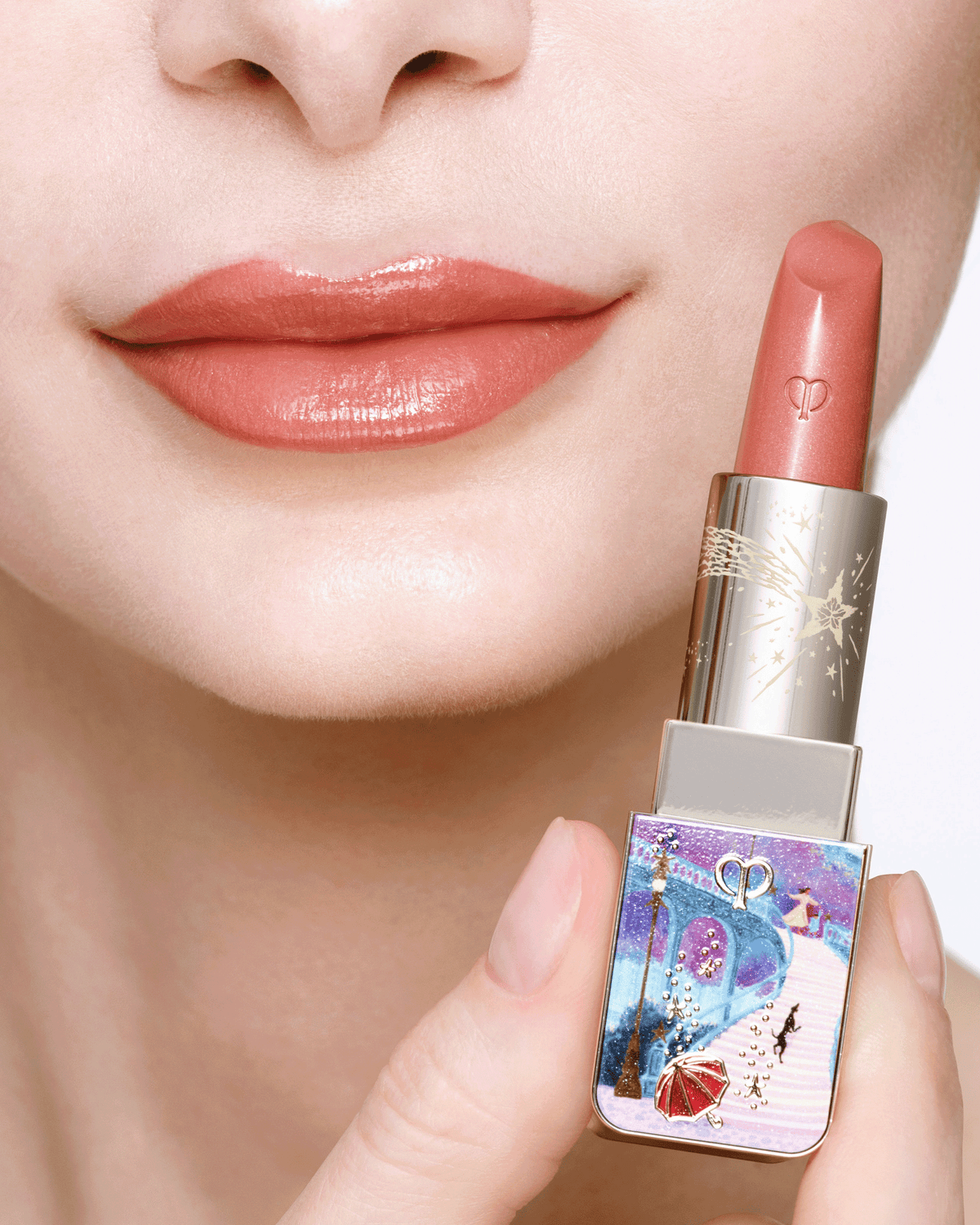 Model with fair skin wearing luminous rose-pink lipstick and holding the Clé de Peau Beauté holiday lipstick with illustrated packaging.