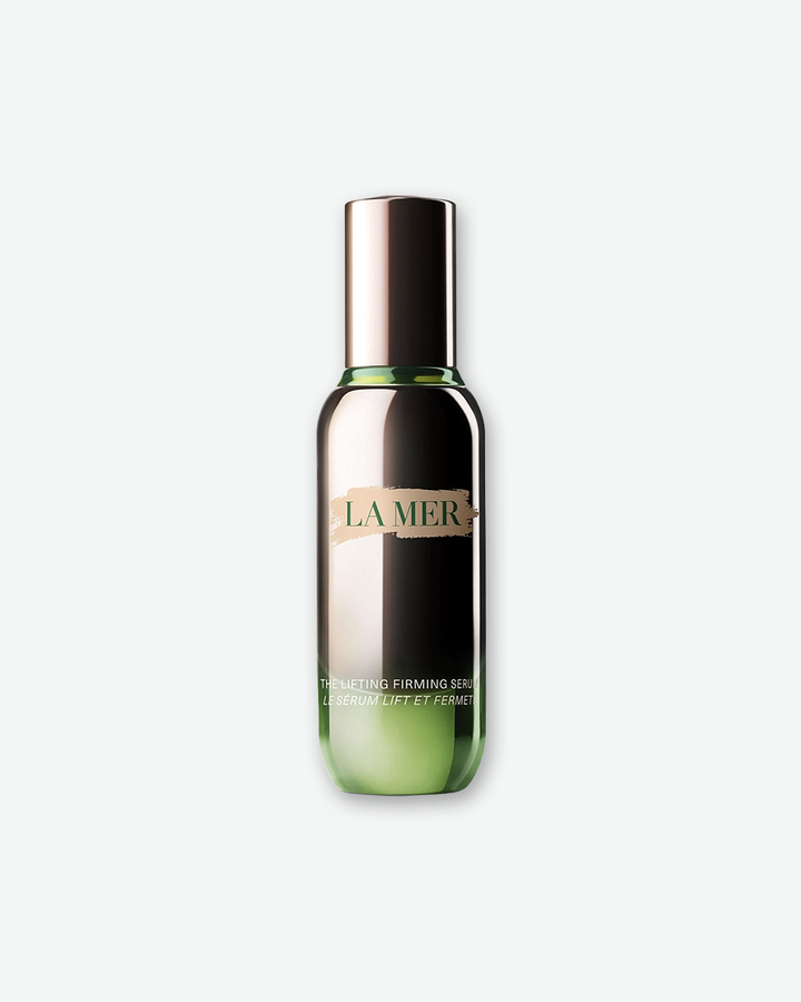 A product shot of a La Mer The Lifting Firming Serum bottle, centered against a plain light gray background. The tall, slender cylindrical bottle is crafted from reflective metallic gunmetal glass with a translucent green base, finished with a matching gunmetal cap. The front displays a gold paint-stroke graphic holding the dark green "LA MER" logo, with white text at the bottom reading "THE LIFTING FIRMING SERUM / LE SÉRUM LIFT ET FERMETÉ".
