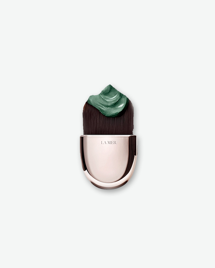 A product shot of a La Mer facial brush, centered against a plain light gray background. The applicator features a smooth, metallic champagne-gold handle. Its dense, dark brown bristles are cut into a wide, flat profile, holding a thick dollop of a dark emerald-green cream mask on top.