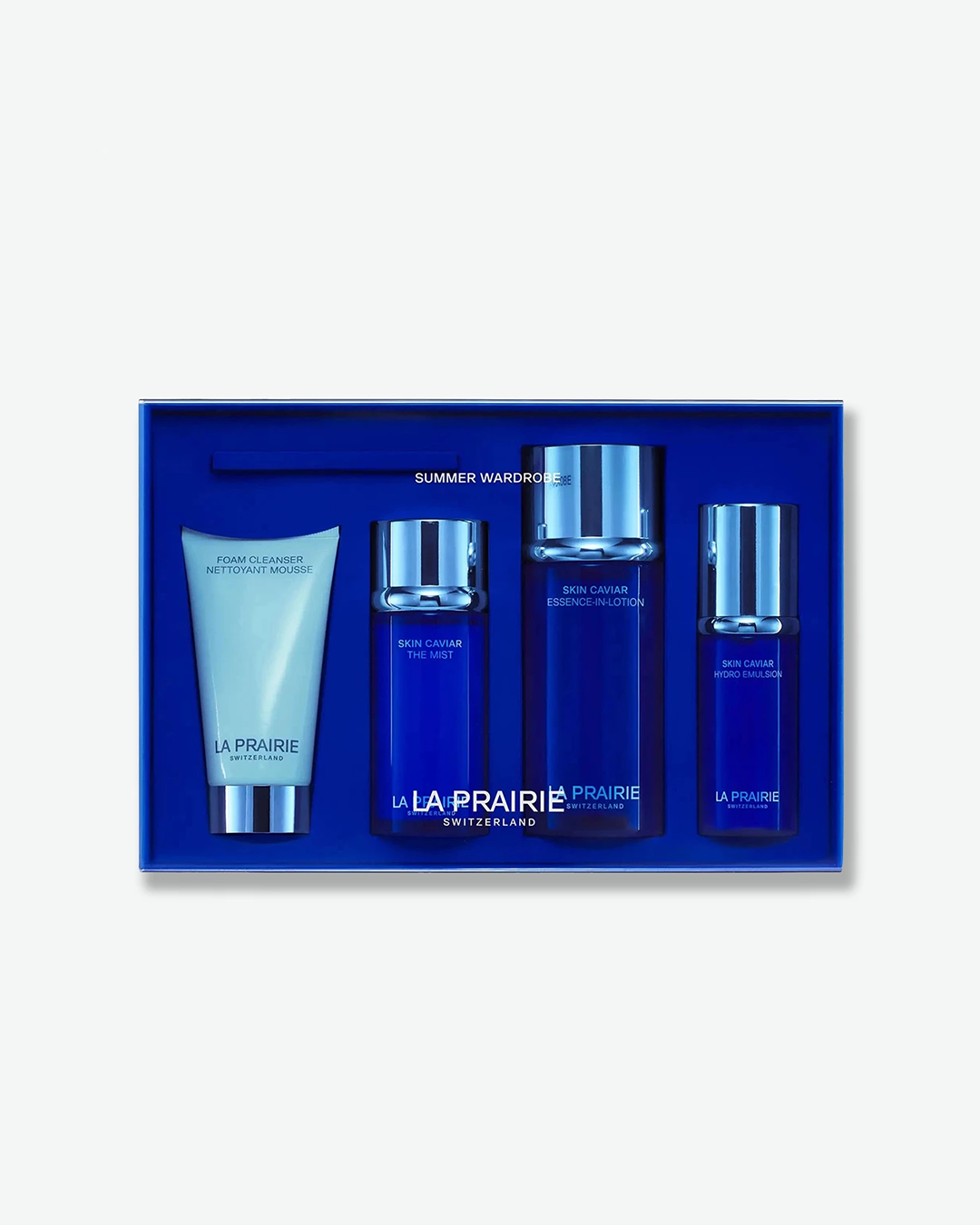 A La Prairie skincare set inside a cobalt blue box, featuring white text overlays that read "SUMMER WARDROBE" and "LA PRAIRIE SWITZERLAND". The set contains a Foam Cleanser tube alongside bottles of Skin Caviar The Mist, Essence-in-Lotion, and Hydro Emulsion.
