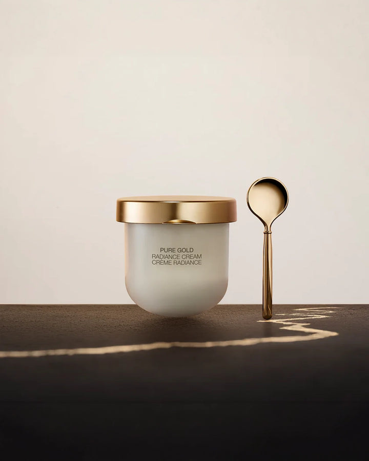 A frosted white glass jar of La Prairie Pure Gold Radiance Cream with a matte gold lid, sitting on a dark surface next to a matching gold applicator spoon.