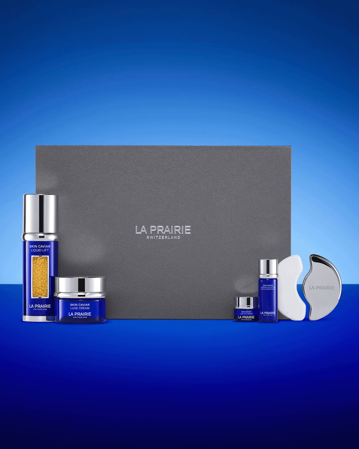 La Prairie Skin Caviar gift set displayed against a signature blue gradient background with the grey presentation box, four blue Skin Caviar products and the silver-and-white facial sculpting tool.