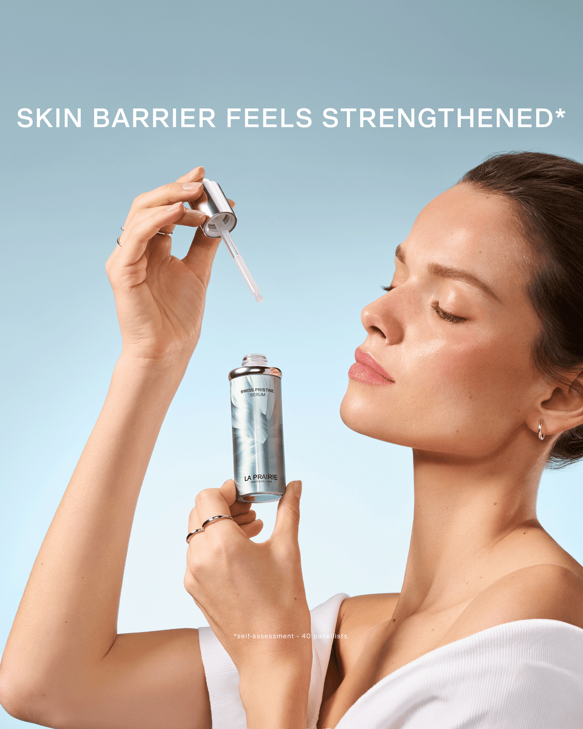 Woman holding La Prairie Swiss Pristine Serum and dropper against a soft blue background with the message “Skin Barrier Feels Strengthened.”