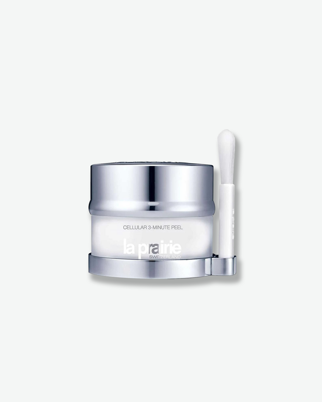 Product shot featuring a La Prairie Cellular 3-Minute Peel jar with its companion application brush. The frosted white glass jar is nestled on a polished silver base that includes a matching vertical holder on the right side for its sleek white applicator brush.