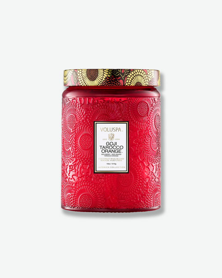 Front view of a Voluspa Goji Tarocco Orange large jar candle against a plain gray background. The candle features a deep red glass jar embossed with a detailed floral motif, a white central label with gold framing, and a decorative tin lid adorned with a black, red, and yellow floral pattern.