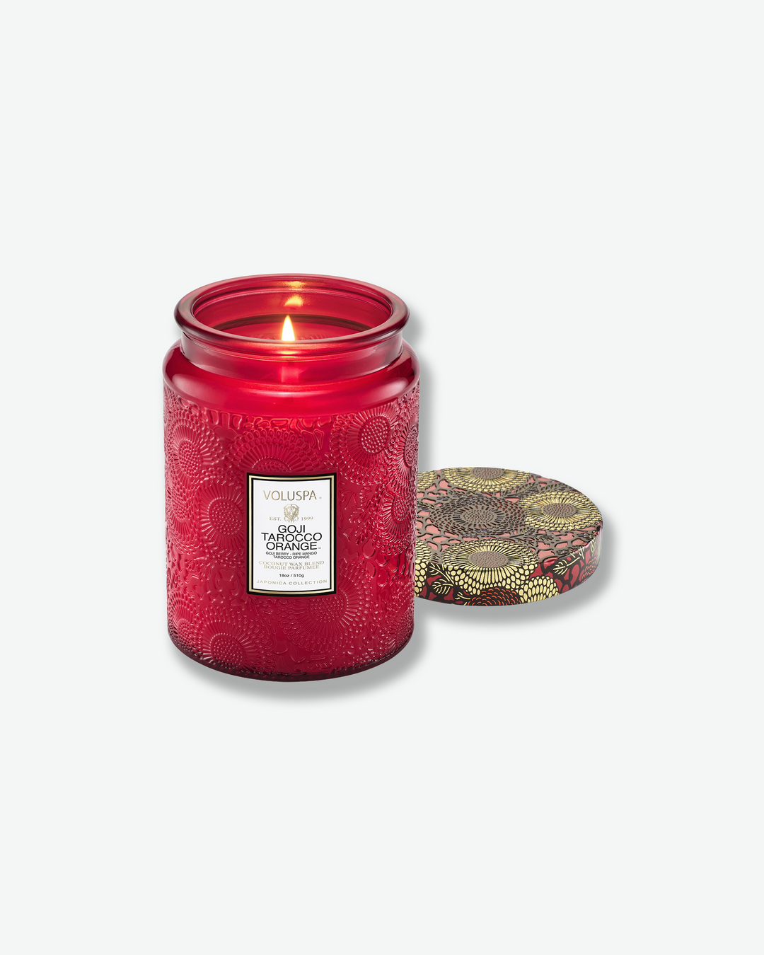 Product shot of a lit Voluspa Goji Tarocco Orange large jar candle. The red embossed glass jar shows a glowing, single-wick flame inside the melted wax. The patterned tin lid is removed and rests flat on its back to the right of the candle.
