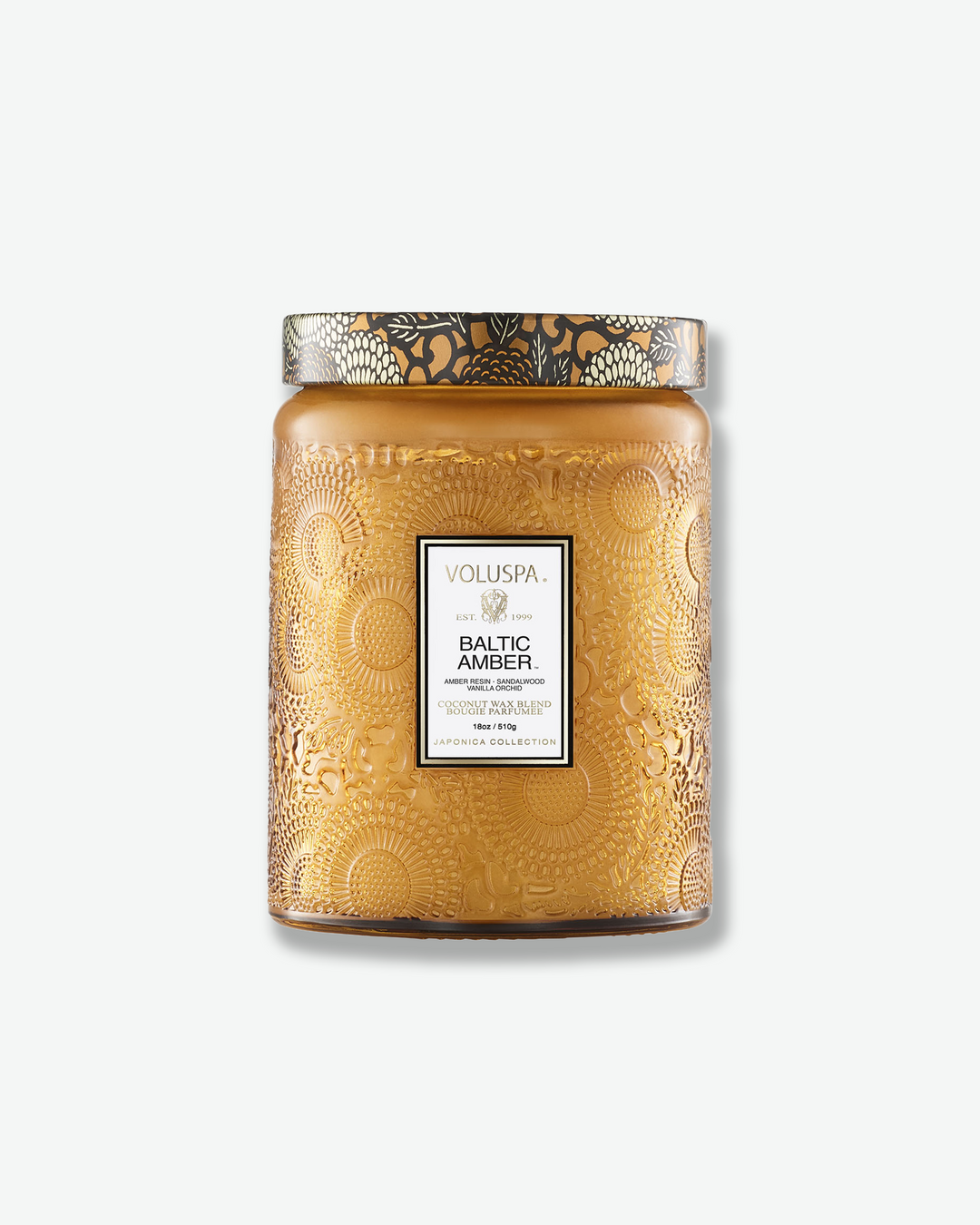 Front view of a Voluspa Baltic Amber large jar candle against a plain gray background. The candle is housed in a warm amber-colored glass jar intricately embossed with floral designs, featuring a central product label and a decorative lid matching the amber and black botanical motif.