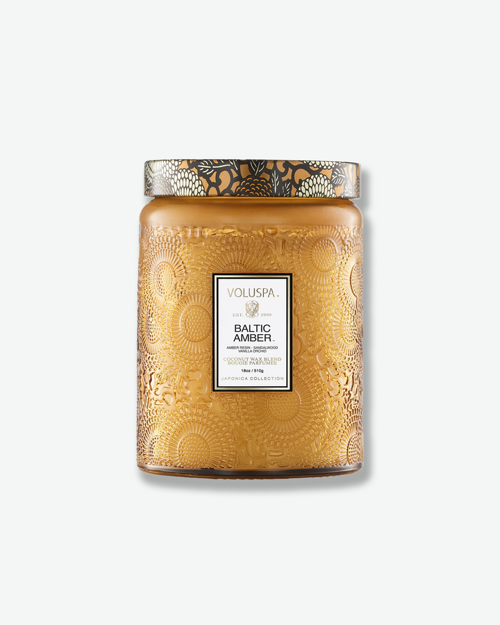 Front view of a Voluspa Baltic Amber large jar candle against a plain gray background. The candle is housed in a warm amber-colored glass jar intricately embossed with floral designs, featuring a central product label and a decorative lid matching the amber and black botanical motif.