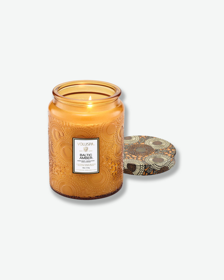 Product shot of a lit Voluspa Baltic Amber large jar candle. The single wick is burning brightly inside the amber glass container, with the patterned amber-and-black tin lid resting face-up next to the jar.