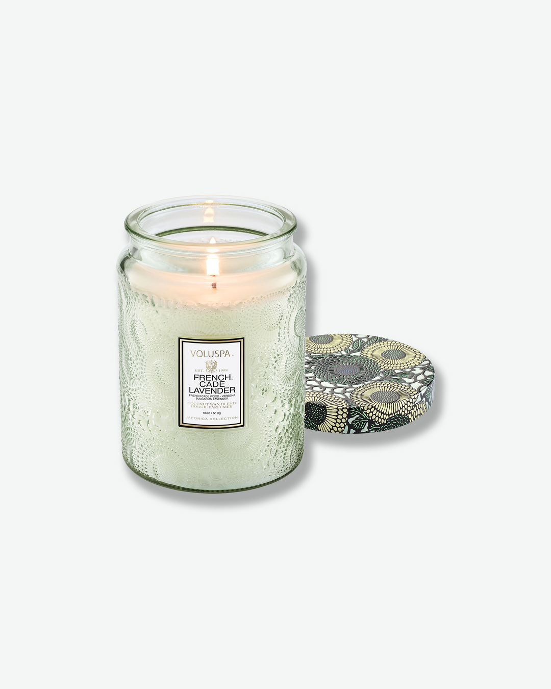 Product shot of a lit Voluspa French Cade Lavender large jar candle. A single flame illuminates the white wax inside the light green embossed jar, with the decorative botanical lid placed flat on the surface next to it.