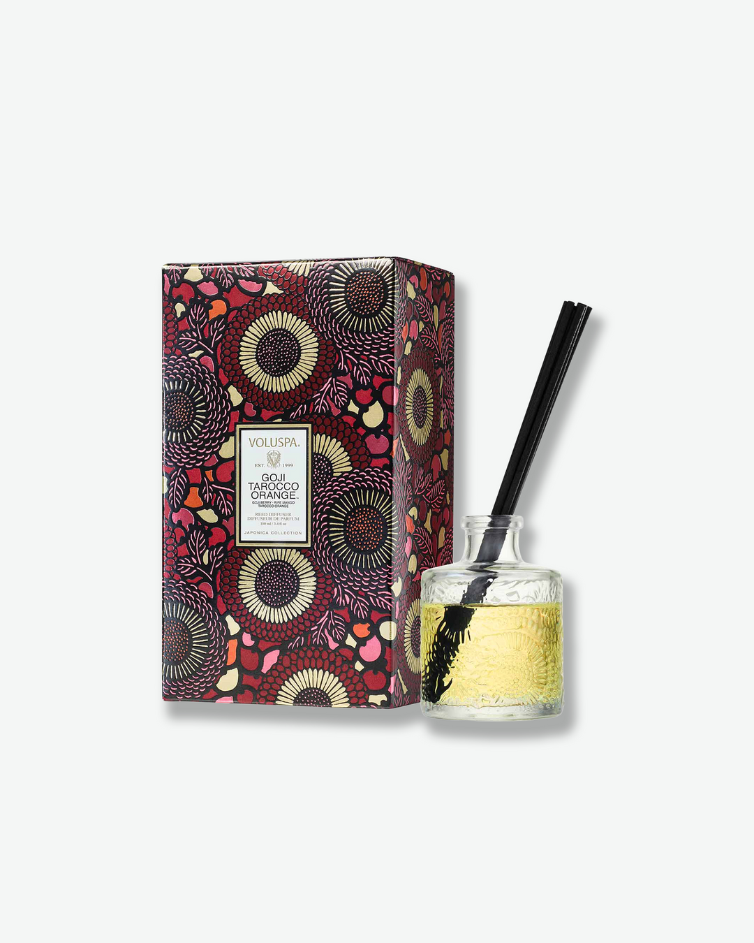 Product shot featuring a Voluspa Goji Tarocco Orange reed diffuser standing next to its decorative packaging box. The clear, embossed glass bottle with a few black reeds sits beside a vertical rectangular gift box adorned with an intricate red, black, and yellow floral pattern.