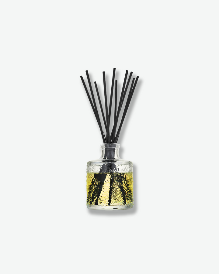 Front view of a Voluspa reed diffuser against a plain gray background. The small, clear glass bottle is embossed with a floral pattern and filled with a light yellow fragrance oil, holding a fan of thick black diffuser reeds extending vertically from the neck.