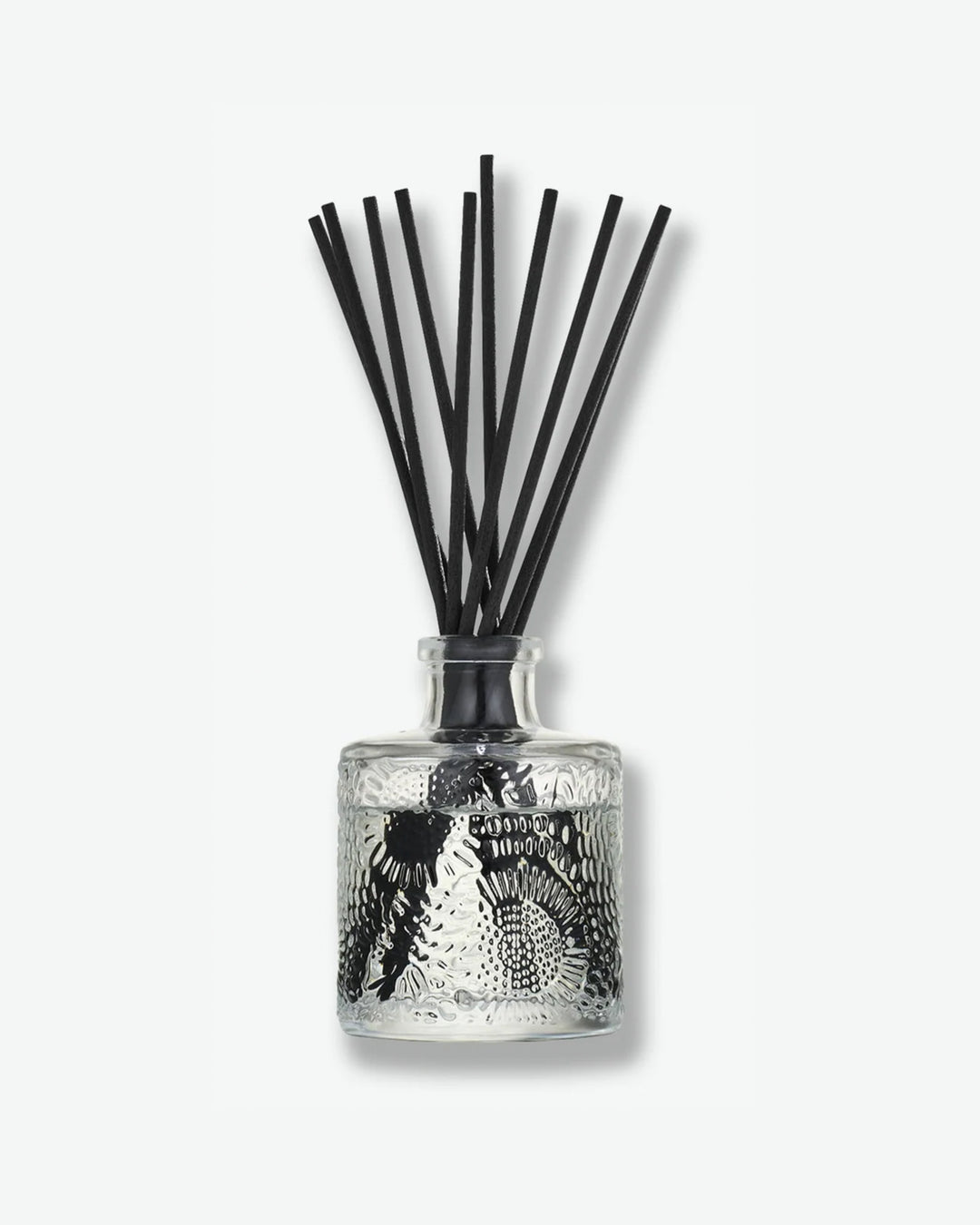 Front view of a Voluspa reed diffuser bottle against a plain gray background. The clear glass vessel is intricately embossed with a floral, swirling pattern and contains clear fragrance fluid, holding a bouquet of dark black diffuser reeds fanning outward from the neck.