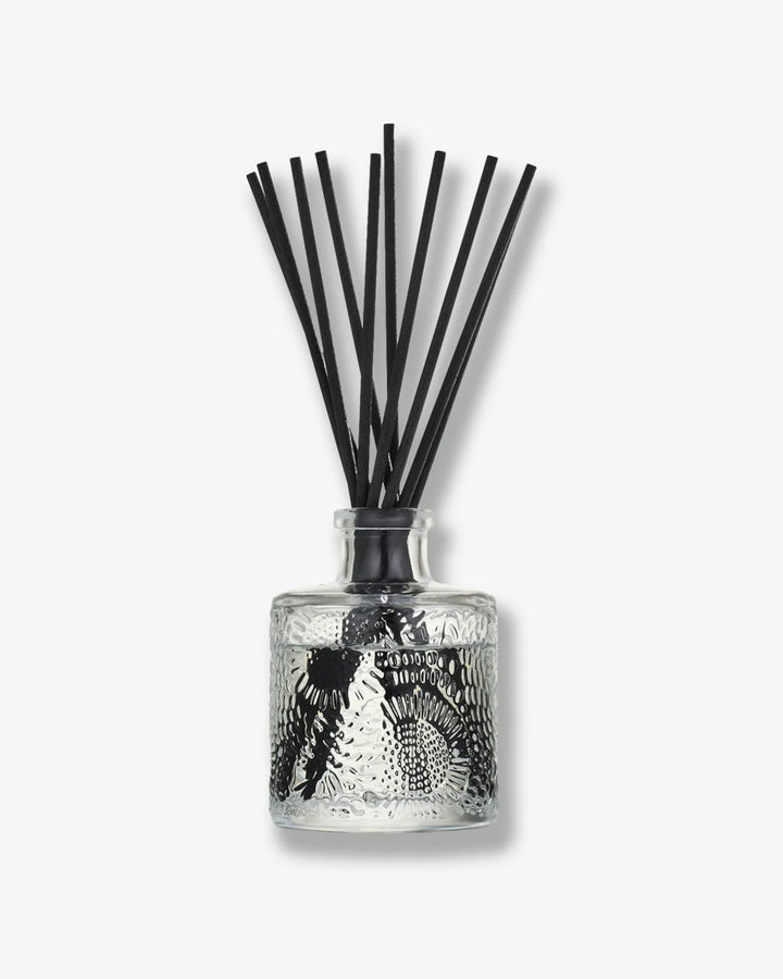 Front view of a Voluspa reed diffuser bottle against a plain gray background. The clear glass vessel is intricately embossed with a floral, swirling pattern and contains clear fragrance fluid, holding a bouquet of dark black diffuser reeds fanning outward from the neck.