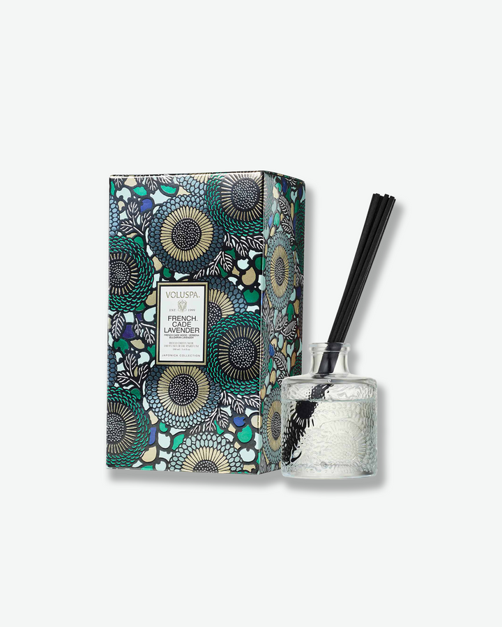 Product shot featuring a Voluspa French Cade Lavender reed diffuser paired with its matching packaging box. The clear, embossed glass diffuser bottle stands to the right of a vertical rectangular gift box adorned with a rich blue, green, and gold botanical design.