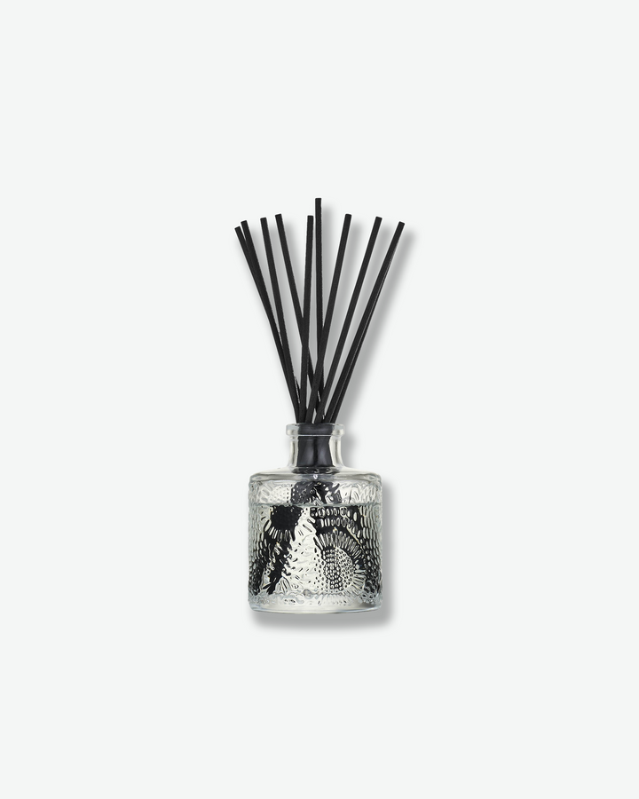 Front view of a Voluspa reed diffuser bottle against a plain gray background. The clear glass vessel is intricately embossed with a floral, swirling pattern and contains clear fragrance fluid, holding a bouquet of dark black diffuser reeds fanning outward from the neck.