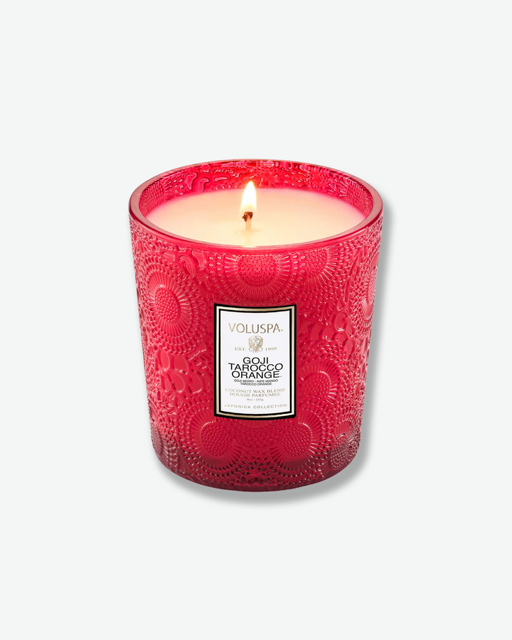Product shot showcasing a Voluspa Goji Tarocco Orange classic textured glass candle next to its square outer box. The red glass candle vessel features a tapered shape and a central white product label, sitting beside a box patterned with the signature red, gold, and black Japonica floral design.